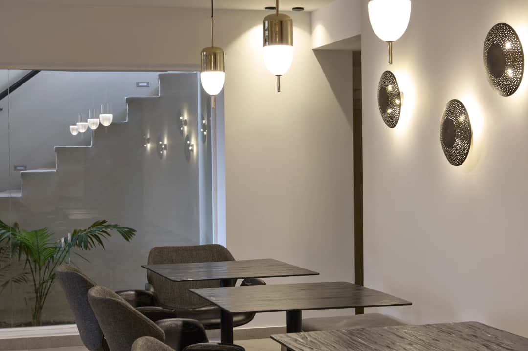 Breakfast room with dark tables and grey chairs under brass pendant lamps, a lit staircase and a palm behind a glass wall