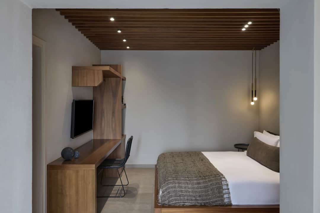 Double bed with a woven throw across from a wooden desk, wall TV and black wire chair, under a slatted wooden ceiling