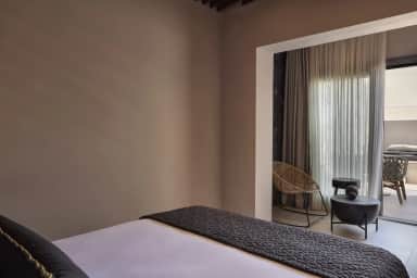 Bed with a charcoal quilted throw facing a curtained nook with a round cane chair, a black side table and a terrace door