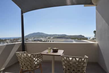 Two woven rope chairs and a wooden table under a shade sail on a balcony over white rooftops, a mountain and the sea beyond