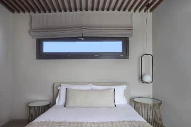 Double bed under a slatted wooden ceiling and a slim window with two grey Roman blinds, gold wire side tables on either side