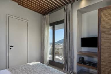 Bedroom with a wall TV above a coffee station and curtains framing a glass door to a sunny balcony with a woven chair