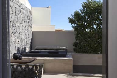 Cushioned daybed on a stone plinth by a volcanic stone wall, a leafy tree in a planter and a woven chair in front