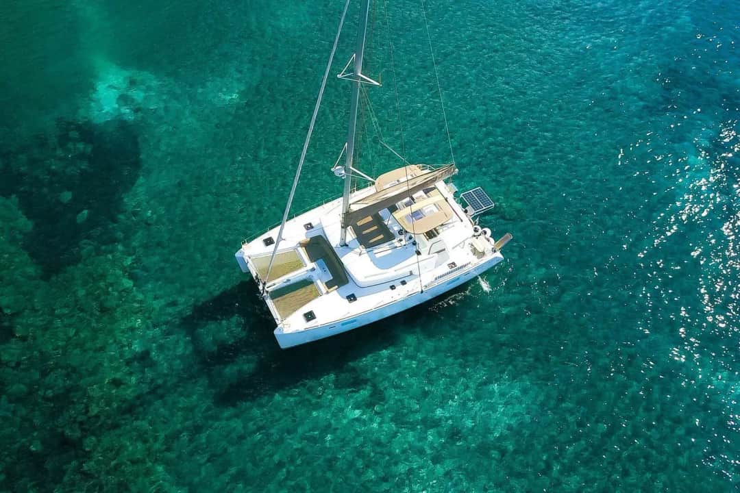 Overhead view of a white sailing catamaran on clear turquoise water, with dark patches and pale sand visible on the seabed