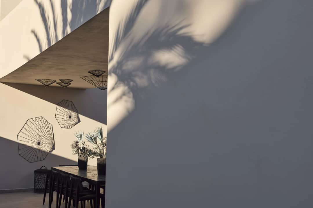 Low sun throwing palm shadows across white walls by a covered dining table with vases of palm leaves and wire ceiling lamps