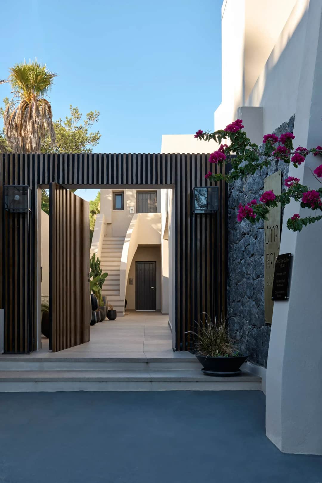 Open wooden gate of Uma Ray Suites framed by bougainvillea, leading to a white courtyard with stairs