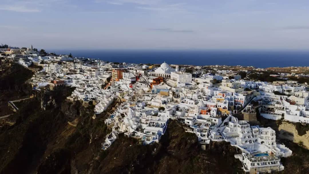 Aerial view of Fira's white houses spilling down dark cliffs around a large domed church, with open sea beyond in low sun