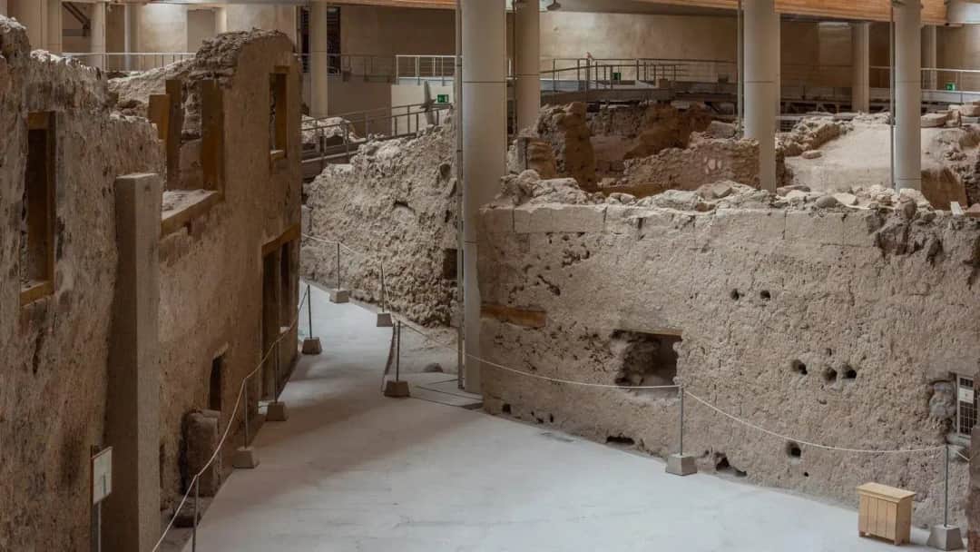 Covered archaeological site with excavated stone buildings, window openings and roped walkways under a roof on white pillars