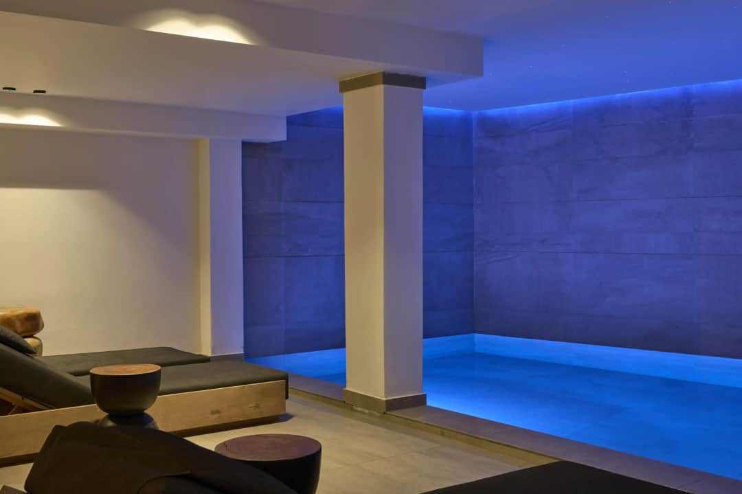Blue-lit indoor thermal pool at Uma Ray Suites seen past cushioned loungers, round wooden side tables and a white pillar