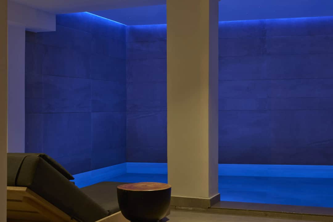 Lounger with a dark cushion and a round wooden table beside the indoor thermal pool, its stone walls washed in blue light