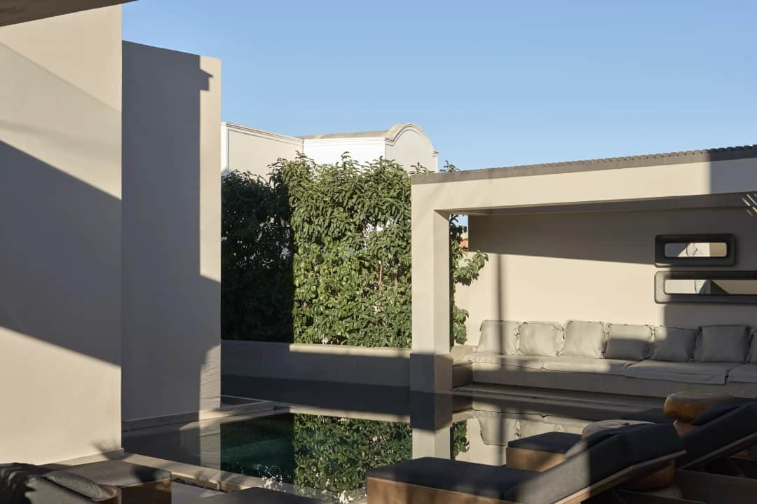 Long shadows over the outdoor pool at Uma Ray Suites, leafy trees mirrored in the water beside a covered sofa lounge