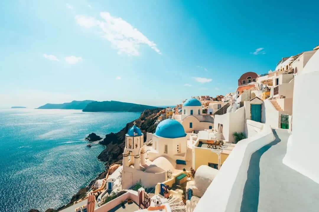 White churches with blue domes and a bell tower in a clifftop village, a whitewashed lane curving above a deep blue sea