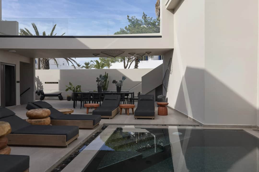 Courtyard with dark-cushioned sunbeds and round wooden stools beside the pool, a covered dining table and palm trees beyond