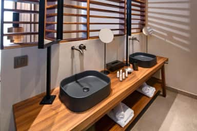 Twin dark basins on a wooden vanity with round mirrors and rolled towels on the open shelf below