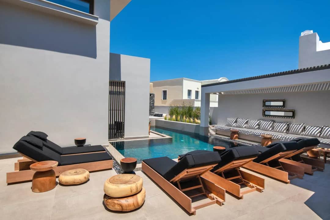 Outdoor pool at Uma Ray Suites in Fira, Santorini, with black-cushioned sunbeds and a shaded lounge