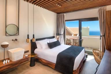 Double bed on a live-edge wooden frame, a stone basin with a round mirror and a balcony with a sea view