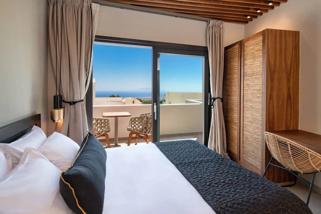 Deluxe Double Room bed facing glass doors to a balcony with two chairs and a view of the sea