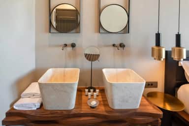 Two white stone basins on a wooden vanity with round mirrors, beside the bed and brass pendant lights