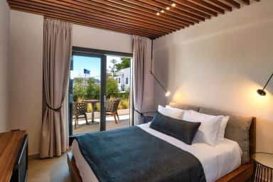 Double bed with wall lamps and glass doors to a small terrace with two woven chairs and plants