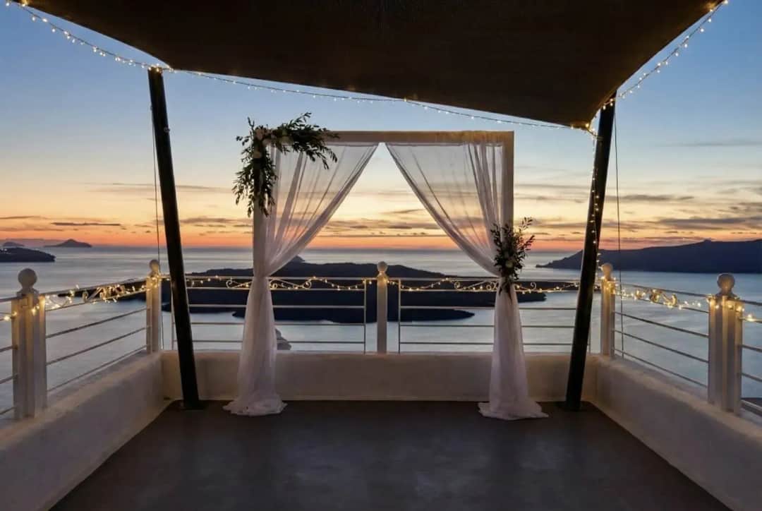 Wedding arch in sheer white drapes and greenery on a terrace strung with fairy lights, facing the sea and islands at sunset