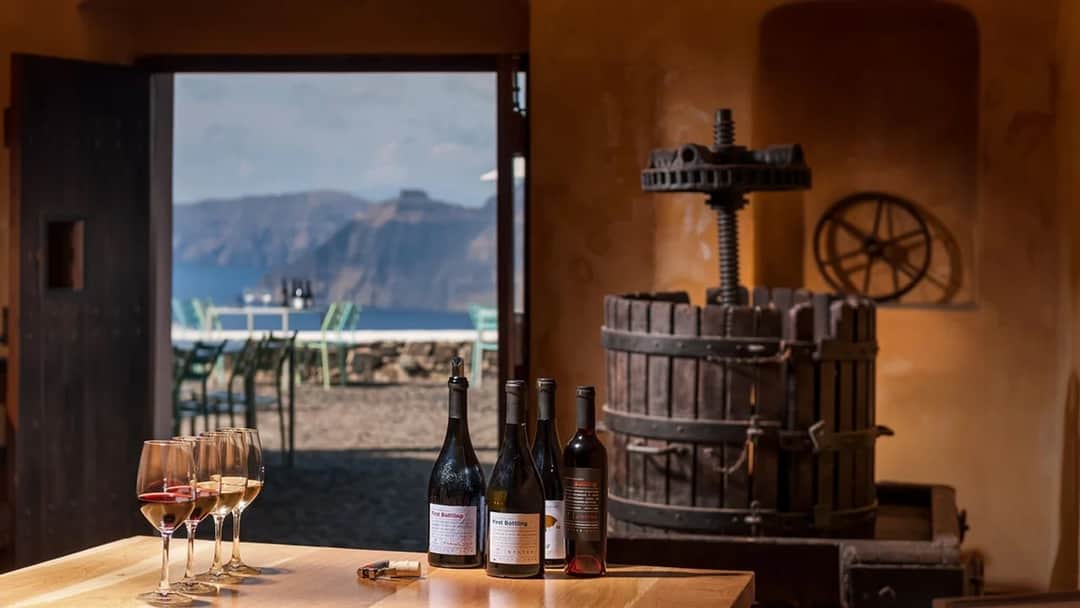 Glasses of wine and bottles on a tasting table beside an old wooden wine press, an open door framing sea cliffs beyond